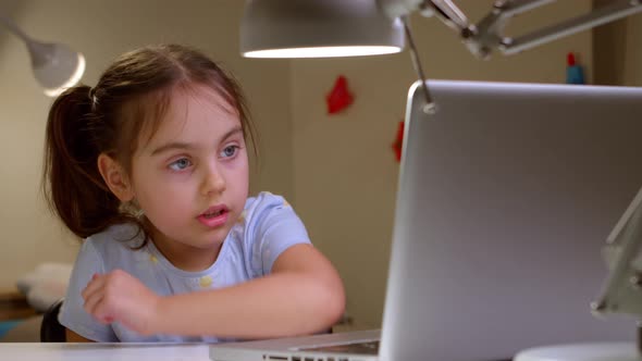 Cute Preschool Girl is Sitting at Her Desk and Looking at Her Laptop