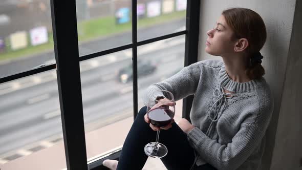 A young woman with a glass of wine sits on the windowsill of a city apartment