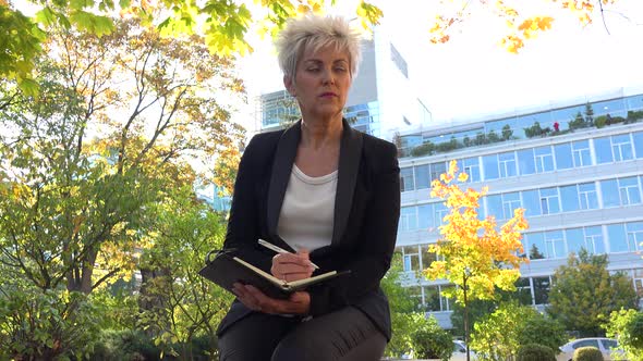 A Middle-aged Businesswoman Sits on a Bench in a Park and Writes Notes Into a Notebook