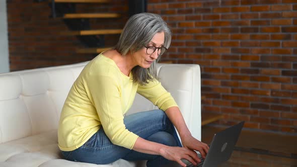 Beautiful Positive Middleaged Woman Sitting on the White Couch and Using Laptop