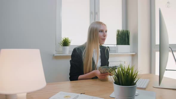 Woman Celebrating Success in Office. Elegant Blond Female Sitting at Workplace Holding in Hands