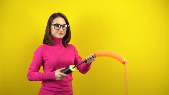 A Young Woman Inflates a Long Orange Balloon with a Pump on a Yellow Background. Girl in a Pink