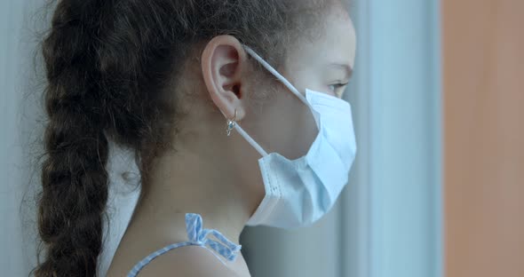 Portrait of a Cute Little Child or Little Girl in a Protective Medical Mask Looks Out the Window