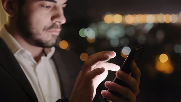 Closeup Serious Young Businessman Using His Phone at Night By Window With Bokeh Lights Background