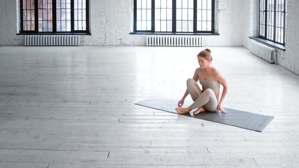 Positive slim young woman in light grey tracksuit comes to mat and sits down in lotus pose