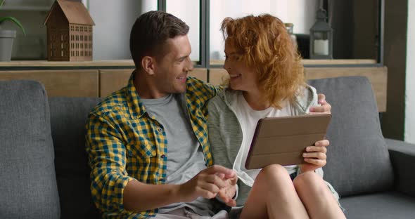 Happy Young Couple Sitting on Sofa, Using Tablet Computer