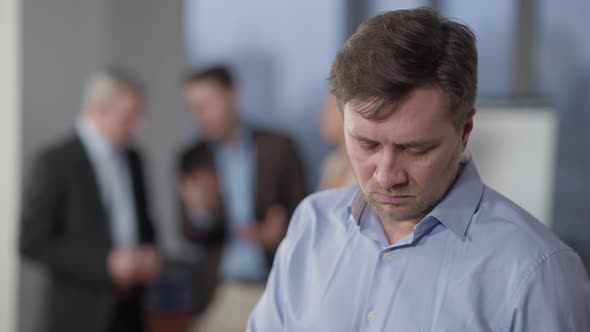 Angry Man Swiping Smartphone or Tablet in Office with Blurred People Chatting at Background