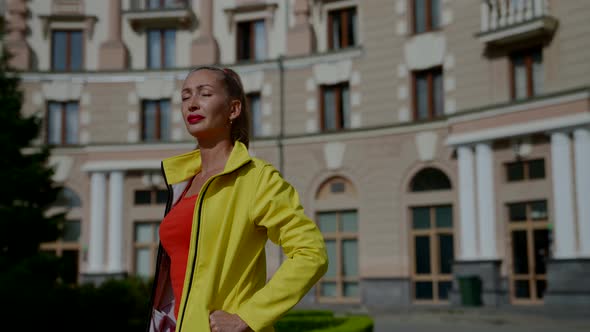 a Woman in a Red Tshirt and a Yellow Jacket Walks Against the Background of a House with Columns
