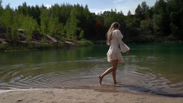 One Young Woman in a White Dress Is Walking and Wetting Her Feet on the Shore of a Wild Forest Lake