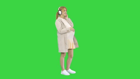 Young Attractive Pregnant Woman Listening To Music in Headphones on a Green Screen, Chroma Key.