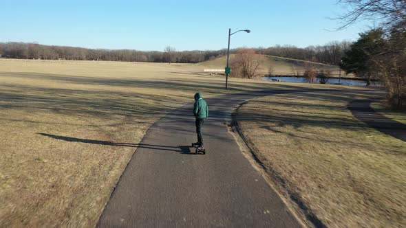 An aerial tracking of a man on an electric skateboard in an empty park on a sunny day. The drone fol