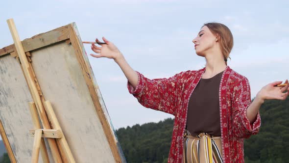 Young Girl Artist Paints Landscape. Girl Stands On Hill And Holds Brush. She Makes Movements Like An