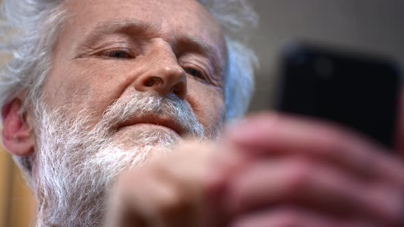 A Grayhaired Old Man with a Beard Holds a Smartphone in His Hand Uses It