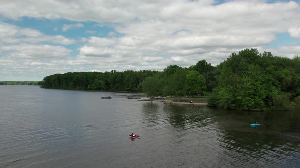 Aerial drone shot of tourists kayaking over a lake beside a park on a cloudy day.