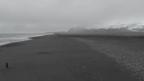 Aerial shot of a woman running along a black sand beach with panning towards the mountains.