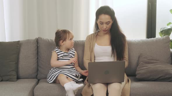 Calm Young Mother Sit on Couch Working on Laptop Little Girl Pulls Her Hand Wants to Play