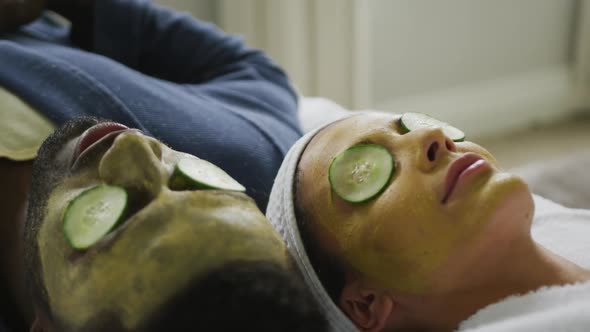 Happy diverse couple lying with masks and pieces of cucumber on eyes in bathroom