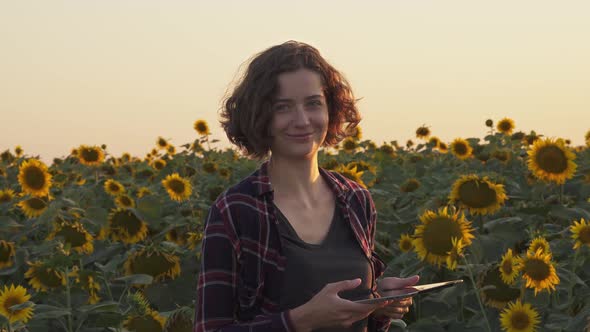 Farmer Woman Stands In Field Blooming Sunflowers, Smiles At Sunset. Portrait Of Joyful, Contented