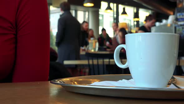 A Woman Sits at a Table with a Cup in a Cafe - Closeup on the Cup, the Interior in the Background