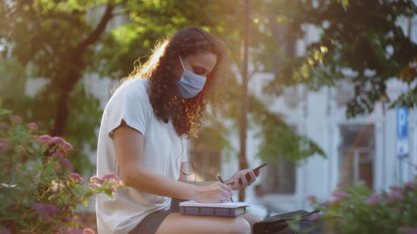 Teen girl college student wearing medical mask and headphones hold mobile phone distance learning.