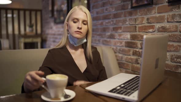 Young Busy Woman in a Medical Mask Works at a Laptop and Drinks Delicious Coffee in a Cozy