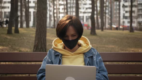 Woman in mask using laptop computer sitting in spring park, working online