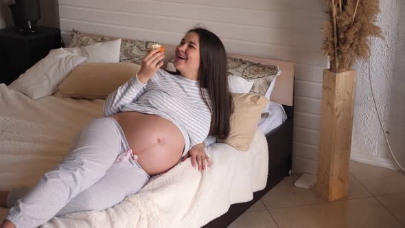 Beautiful Young Pregnant Woman Eating Cake in Bed at Home and Smiling