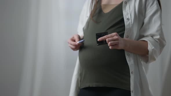 Pregnant Unrecognizable Caucasian Woman Holding Pregnancy Test and Condom Standing Indoors