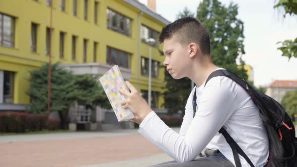 A Caucasian Teenage Boy Reads a Book  Side View  a School in the Background