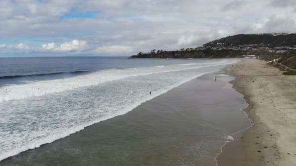 A drone flies over crashing ocean waves at the beach as surfers paddle out to surf.