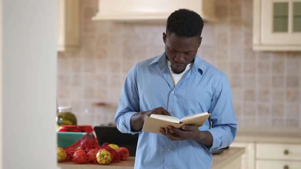 Focused Young African American Man Reading Recipe Looking Back at Vegetables on Table in Kitchen