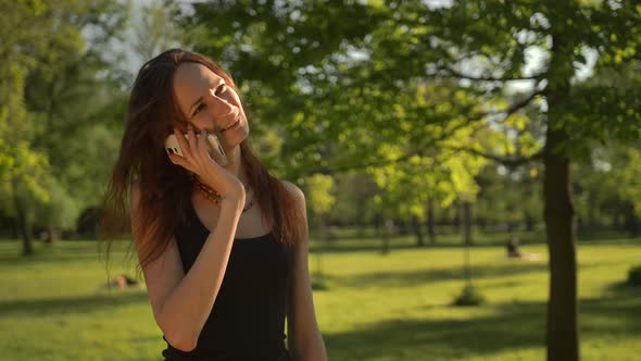 Smiling Young Woman Stand at the Park Outdoors Talk By Phone