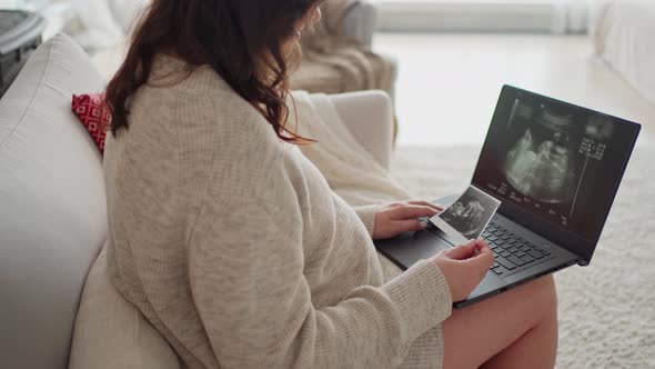 a Young Pregnant Woman Sits on a Sofa and Watches a Video of an Ultrasound of Her Unborn Child on a