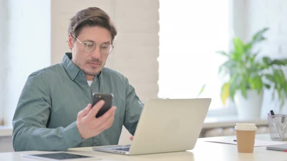 Middle Aged Man with Laptop Using Smartphone in Office
