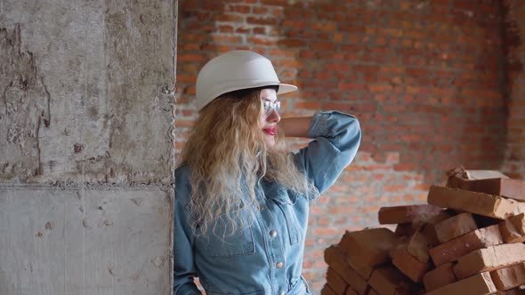 A Young Woman in a Work Helmet and Denim Clothes and Goggles Stands at the Construction Site
