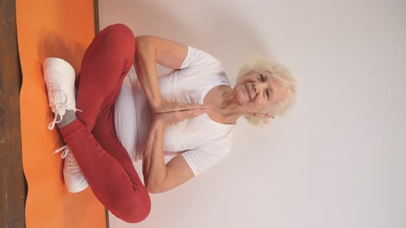 an Elderly Woman in Sports Clothes in the Lotus Position Sits at Home on the Mat of Morning Exercise