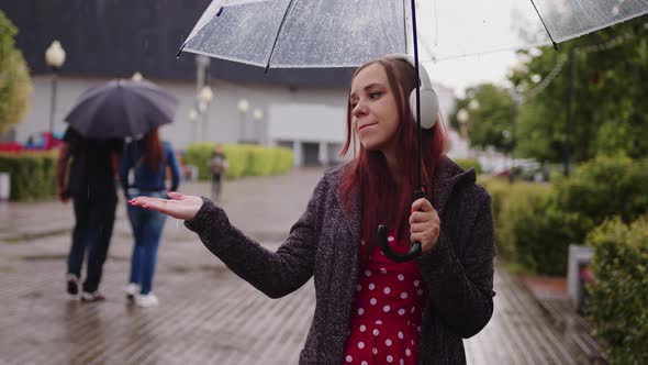 Young Beautiful Woman in Headphones Listening to Music Standing with Transparent Umbrella in Rain on