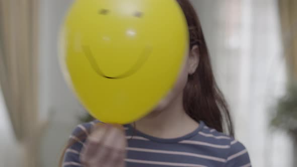 A Pretty Girl Hiding Behind Yellow Balloon with a Painted Happy Face on It Then Puts It Down