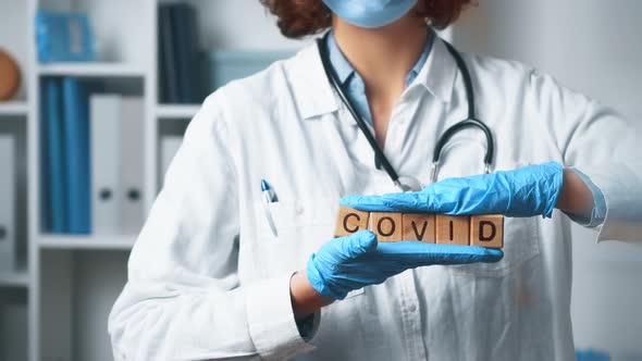 The Word Covid Is Held In Hands. A Doctor In A Medical Uniform Stands In A Hospital Medical Office