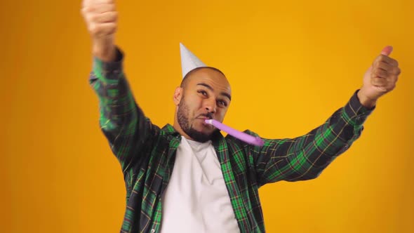 Happy African American Male in Party Cone Blowing Party Blower Against Yellow Background