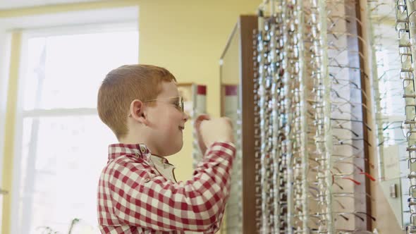 A Redhaired Boy in a Plaid Red and White Shirt Tries on Sunglasses Near a Rack with Lens Frames and