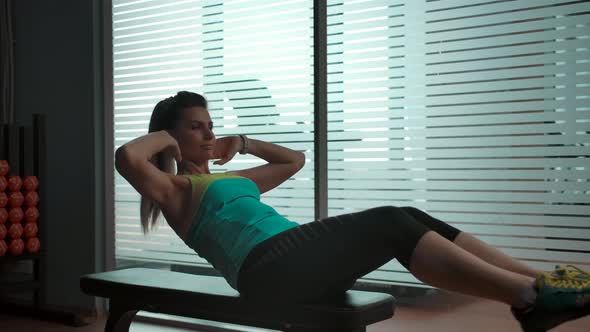 The Girl Does Abs Exercises on a Bench in the Gym Next to a Large Window