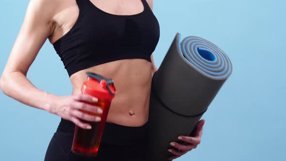 Young Caucasian woman in black sports top holding up a yoga mat and a pink water bottle