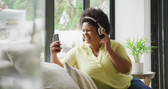 Video of happy plus size african american woman with headphones on sofa, having video call
