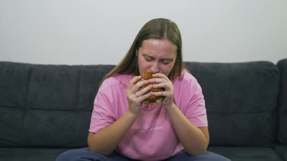 Plump Young Woman with Blue Eyes and Pink Tshirt is Sitting on Gray Sofa and Eating Big Fat Burger