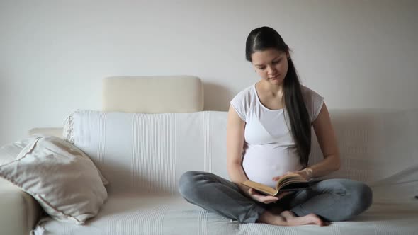 Young Pregnant Woman Sitting on White Sofa in Home Room and Reading the Book