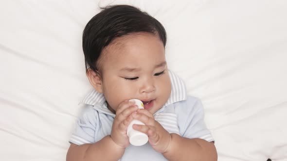 Asian Baby Boy Biting On A Bottle Teether While Lying In Bed. overhead