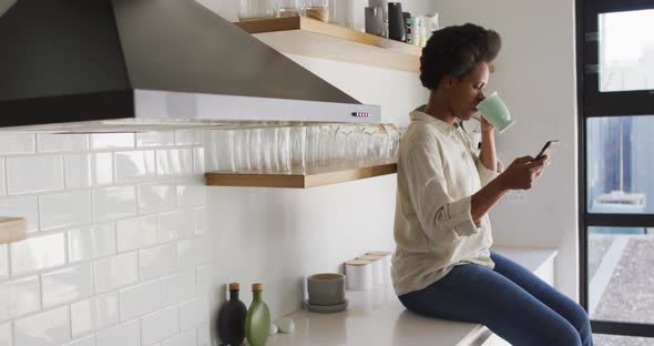 Happy african american woman drinking coffee and using smartphone in kitchen