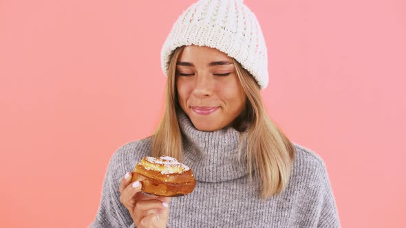 Female in Warm Sweater and Hat is Holding a Bun Sprinkled with Icing Sugar and Smiling While Posing