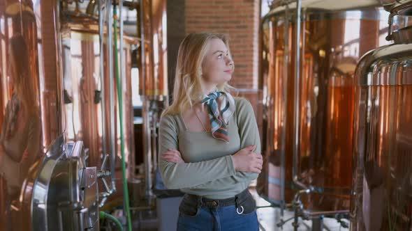 Slim Confident Smiling Woman Standing in Beer Brewery at Large Tanks Looking Around